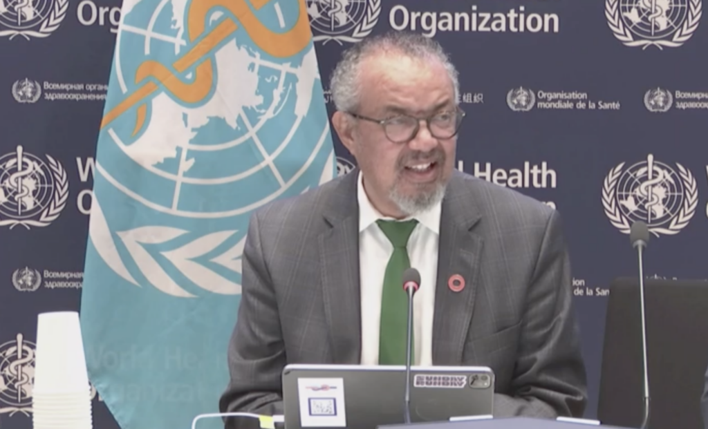 Man in a gray suit and green tie speaks at a WHO press conference, with a UN backdrop and microphone in front of him.