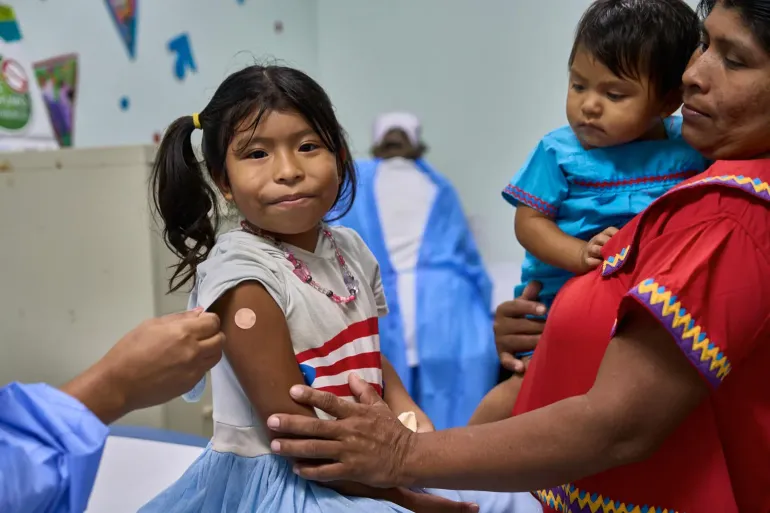 Girl seated as a nurse administers a vaccine to her upper arm in a clinic; caregiver holds a young child nearby, calm atmosphere.