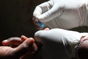 Gloved hands performing a finger-prick with a blue lancet to collect a blood sample.