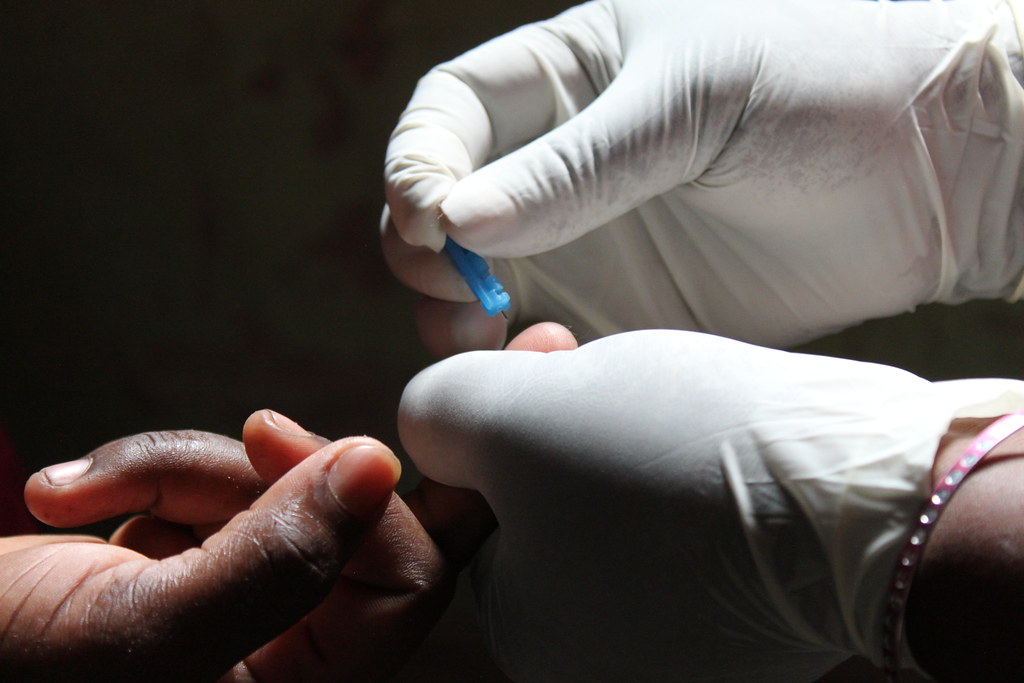 Gloved hands performing a finger-prick with a blue lancet to collect a blood sample.