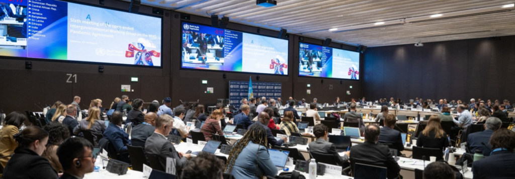 Delegates in a large international conference hall watching multiple screens announcing a pandemic agreement meeting at the UN/WHO event.