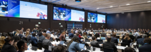 Delegates in a large international conference hall watching multiple screens announcing a pandemic agreement meeting at the UN/WHO event.