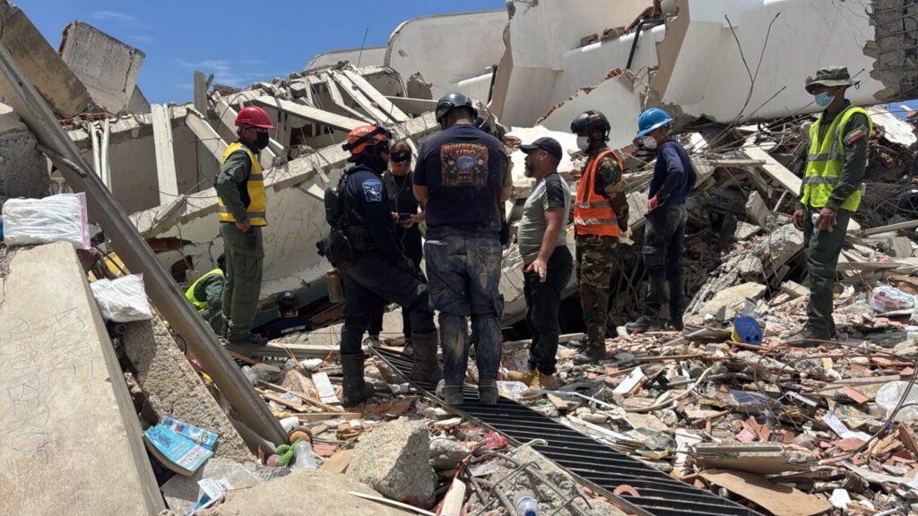 Emergency responders wearing helmets and high-visibility vests stand on a concrete-collapse site, surveying rubble and debris amid a collapsed building.