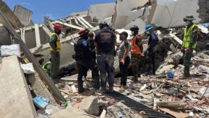 Emergency responders wearing helmets and high-visibility vests stand on a concrete-collapse site, surveying rubble and debris amid a collapsed building.