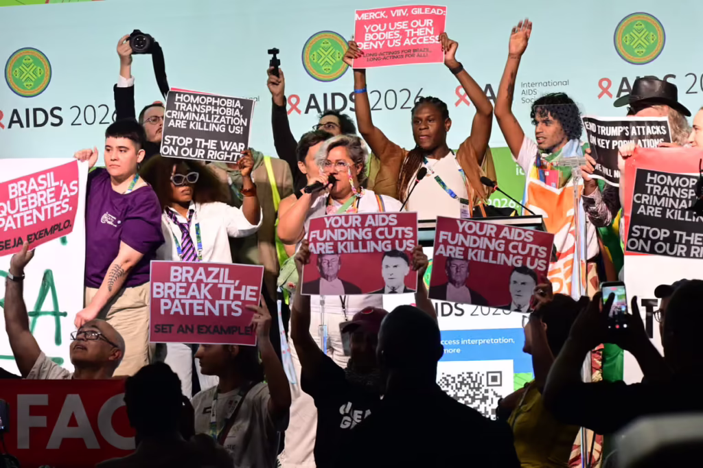 Diverse group on stage at AIDS 2026 rally, holding pink protest signs about funding cuts and patents.