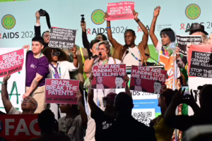 Diverse group on stage at AIDS 2026 rally, holding pink protest signs about funding cuts and patents.