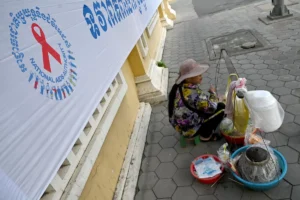 Women selling street snacks on a sidewalk next to a large banner with a red ribbon for National AIDS charity, wearing a hat and floral jacket.