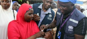 Mother in a red hijab holds her baby as a WHO health worker vaccinates the infant.