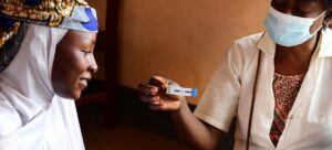 Healthcare worker wearing a mask holds a rapid diagnostic test strip for a seated woman wearing a headscarf.